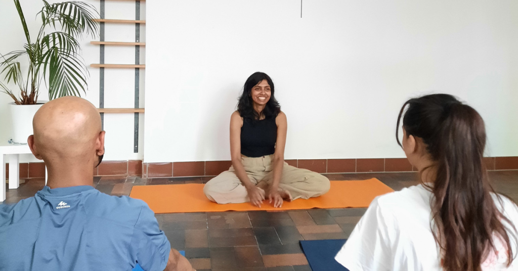 Eine Frau sitzt auf einer Yogamatte und lächelt zwei Teilnehmende, die auch auf einer Yogamatte sitzen an.